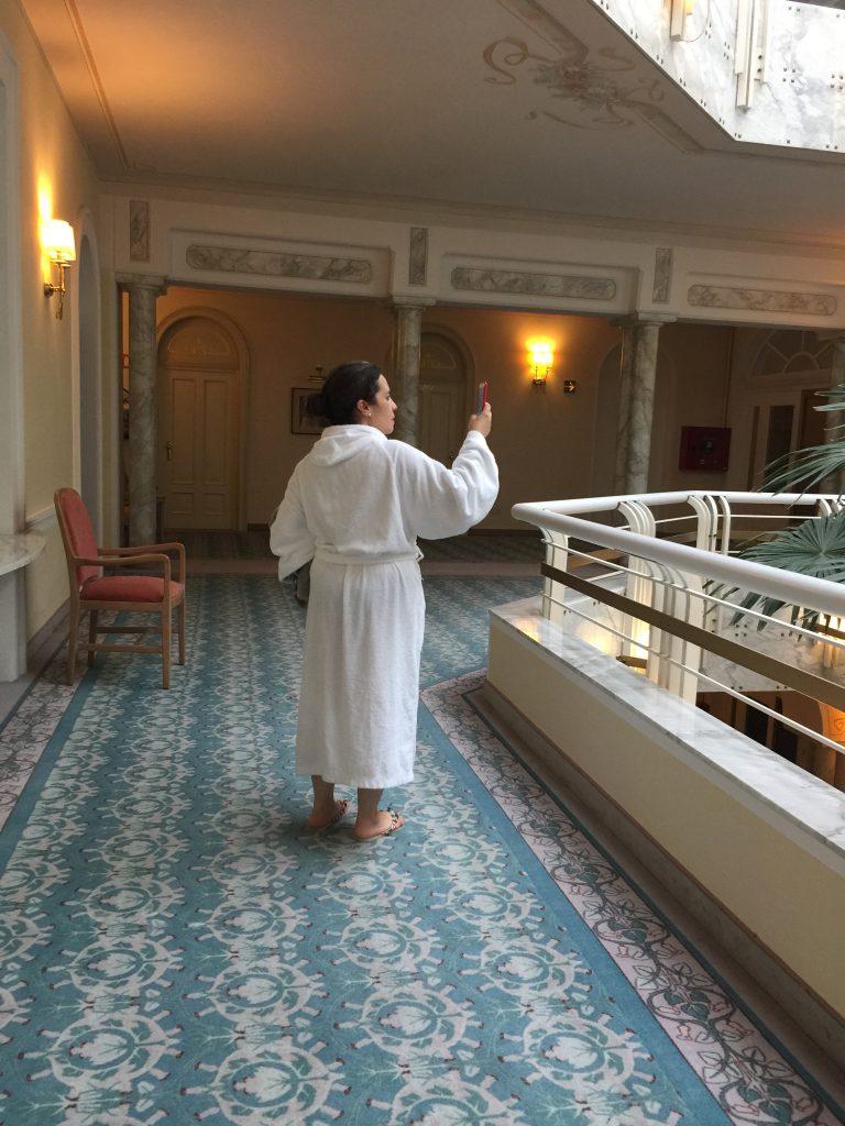 Chiara Carolei indossa un accappatoio bianco pronta per recarsi nella zona termale a rilassarsi. Con il suo smartphone scatta qualche foto e viene fotografata senza che se ne accorga lungo i corridoi dell'hotel